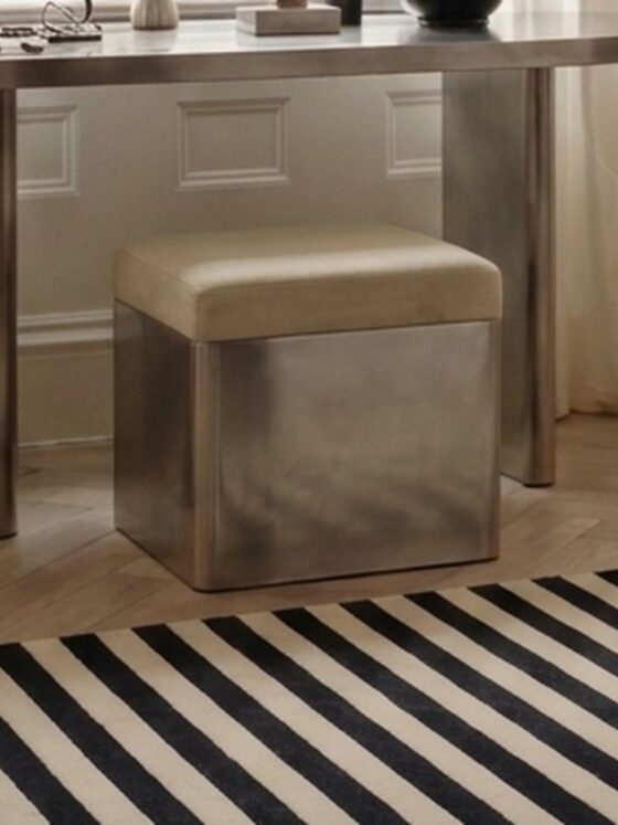 The Kasanai stainless steel stool tucked beneath a stainless steel dressing table, in a calm bedroom with a striped rug.
